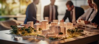 Im Hintergrund sieht man eine Diskussionsrunde von fünf Architekten und im Vordergrund steht das Modell einer Wohnanlage